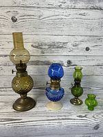 Front view of three colored hobnail glass miniature oil lamps in amber, blue, and green, with varying heights and metal burners.
