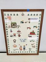 Full view of framed cross-stitch depicting various small rural and domestic motifs with phrase and border of trees.