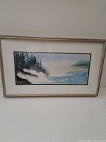 Framed watercolour coastal landscape painting with rocks, waves, and trees.
