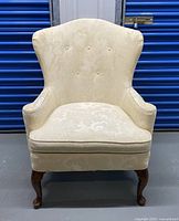 Front view of cream fabric upholstered accent chair showing wingback style with button tufting and wooden cabriole legs.