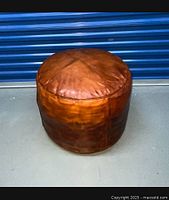 Front view of a brown leather round ottoman with visible texture and creases, set on a concrete floor against a blue metal shutter background.