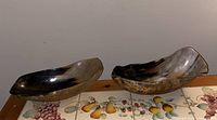 Two polished horn bowls placed side by side on a surface with a decorative fruit motif underneath. The bowls show natural horn coloration with glossy finish.