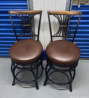 Front view of two swivel bar stools showing brown round cushions, metal frames, decorative curved backs with wooden top rail.