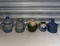Five assorted pottery items arranged side by side: covered jar, two pitchers, small container, and blue bottle.