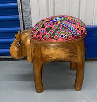 Side angle view of the wooden elephant-shaped foot stool ottoman showing the carved elephant features and colorful embroidered cushion on top.