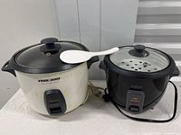 Front view of two electric rice cookers side by side with a white rice spoon on the Back & Decker cooker.
