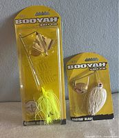 Front view of two Booyah fishing lures in original yellow factory packaging, one larger spinnerbait with yellow skirt and two gold blades, one smaller blade bait spinner with white skirt and one gold blade.