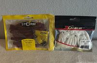 Two packs of sealed soft plastic fishing bait, one Storm Rattle Ribbon Tail Worm in purple with bonus rigging kit, and one pack of white Xcalibur soft plastic grubs.
