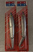 Two new Rebel brand fishing baits in original packaging, silver with treble hooks, displayed on carpet.