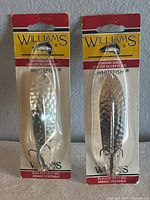 Two Williams Whitefish fishing lures in original packaging, hammered metallic finish, with treble hooks visible.