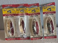 Four Williams WablerLite fishing lures in original factory packaging, showing both gold and silver plated finishes and clear sealed packaging.