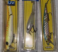 Six Storm fishing baits in factory packaging, various colors including orange/white UV bright, translucent gray, and shiny pink reflective scale.