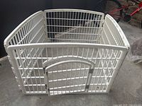 White metal dog pen shown from an angle, display of gate with rounded top and parallel bars.