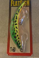 Flatfish T-60 fishing lure in original unopened factory packaging, showing green/yellow body with black spots and two metal treble hooks.