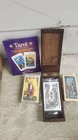 Photo showing three complete tarot decks, one deck with visible 'The Magician' card, a purple tarot guide book and an open wooden storage box.