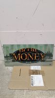 Front view of rectangular slot machine glass panel with 'IN THE MONEY' text and forest design background laid on cardboard against wall.