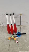 Photo showing set of three red and white juggling clubs standing upright, five colorful juggling balls on the floor, blue diabolo with white string and two black diabolo sticks, plus a black juggling stick with beige rubber top against a white wall.