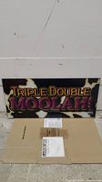 Photo of rectangular plastic slot machine topper with 'TRIPLE DOUBLE MOOLAH!' text and cowhide background, resting against wall and placed on cardboard.