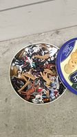 Top-down view of a round tin box filled with mixed plastic animal and vehicle figures, varied colors and wear visible.
