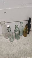 Full view of six vintage glass bottles of various shapes, sizes, and colors arranged on a floor against a white wall.