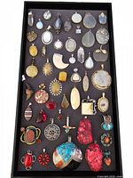 Top-down view of a tray with 50 assorted gemstone pendants of various shapes and sizes laid out on black background