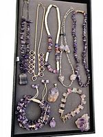 Top-down view of all 10 amethyst jewelry pieces and raw stone displayed on dark tray.