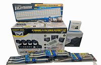 Photo shows complete set of Night Owl 4 channel H.264 video security kit box with 4 cameras and DVR, three Alpena Max LED light strips, Bell+Howell LED Light Bar box, projector box, and IP camera box.