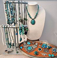 Overall display showing multiple hanging bead necklaces on T-bar, bust with green bead pendant necklace, tray with bib necklace and assorted pieces.