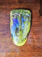 Close-up front view of iridescent glass face sculpture showing blue and yellow hues and detailed molded facial features.