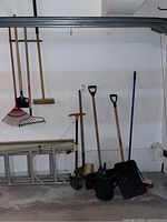 Photo showing full set of garden tools leaning against a wall in garage including rake, broom, hoe, watering can, shovel, string and snow shovel.