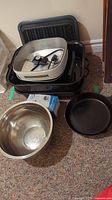 Overall photo showing stacked metal roasting pan, coffee grinder box, electric frying pan, cast iron pan and large mixing bowl on carpet floor