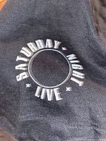 Close up showing a black item of clothing with white 'Saturday Night Live' logo printed in a circular design.