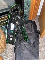 Pair of green frame Big Foot snowshoes with black bindings resting next to a black duffle bag on a carpeted floor near a fireplace.
