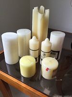 Eight assorted pillar candles displayed on glass surface including a tall irregular ivory pillar candle and smaller white and yellow candles