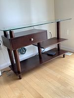 Full view of the dark wood console table with glass top, drawer, and shelves on a light wood floor near a wall and power outlet.