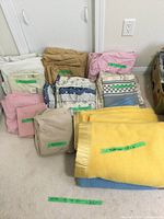 Photo showing folded blankets in yellow and blue wool blend with tags, stacks of king size sheets and pillowcases in beige, pink, white, and patterned fabrics, and some throw pillows.