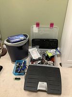Photo showing a Fellowes paper shredder, a plastic box filled with assorted computer cables, a black keyboard, and a black foot stool placed on the floor.