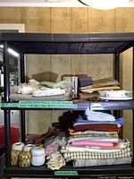 Photo showing two shelves stacked with assorted folded fabrics in various colors and patterns including white lace, pastel green, beige plaid, and pink fabric, along with spools of thread and sewing supplies.