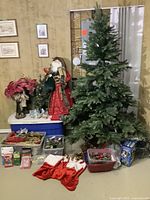Photo shows large artificial Christmas tree, Santa figurine in red clothing, red poinsettia plant, several bins containing various Christmas ornaments, multiple red Christmas stockings laid out in front of tree on floor along with boxes possibly containing lights or decorations.