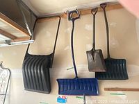 Three snow shovels and one spade hanging on garage wall: large black push-style shovel, blue D-handle snow shovel, green plastic snow shovel, and square metal spade