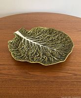 Oval ceramic platter shaped and textured like a green cabbage leaf with white veins, positioned on a wooden table.