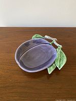 Front view of the ceramic fruit-shaped dish showing plum shape and attached green leaves.