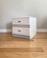 Front view of the white post modern nightstand showing two drawers with gold knob handles and gray accent strip.