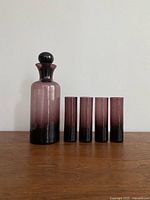 Angled view of vintage purple glass decanter and four matching glasses arranged side by side, showing gradient color and glass finish.
