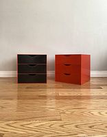 Two modular stacking organizer boxes with three drawers each, one orange and one with orange frame and black drawers, placed side by side on the wooden floor against a white wall.
