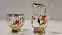 Side view of the small milk pitcher and sugar bowl both featuring gold trim and hand painted red and pink floral designs with gold leaf accents