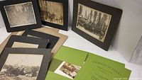 Collection of black and white antique photos mounted on dark and tan boards, depicting logging camps and groups of people in a forest setting.