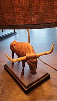 Side angle showing carved wooden bull lamp base with brown textured cloth lampshade attached to wooden base.