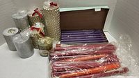 Silver and gold textured pillar candles, small jar candle with gold ribbon, purple taper candles in box, and red/orange taper candles in plastic wrap.