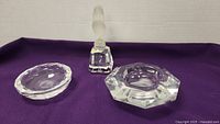 Clear heavy glass items on purple cloth including bird figurine on stand, oval bowl and ashtray.